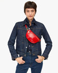 A person in a dark denim jacket and jeans poses confidently with hands in pockets. They have short brown hair and wear the Clare Vivier Petit Fanny F25, an adjustable petite fanny pack, across their chest. The background is plain white.