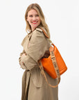 A smiling woman in a beige trench coat carries an orange bag with the Clare Vivier Adjustable Crossbody Strap, featuring bold black text on thick cotton webbing. She stands against a plain white background.
