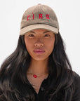 A woman with long black hair models the Clare Vivier Wool CIAO Baseball Hat, featuring tan herringbone wool embroidered with "CIAO" in red. She wears a black shirt and gold necklace with a red heart, facing forward against a plain white background.