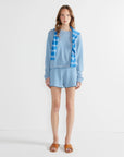 A woman stands against a white wall wearing The Peyton by Kule: a blue and white striped 100% cotton long-sleeve shirt, matching shorts, a striped knit sweater over her shoulders, and brown sandals.