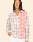 A woman with straight dark hair wears the Xirena Phillipa Shirt—a 100% cotton, collared, long-sleeve button-up in light purple and pink plaid—with white pants, standing against a plain beige background.