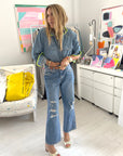 In a vibrant, art-filled room, a woman wears the Pia Denim Western Shirt by Kerri Rosenthal with distressed jeans and open-toe heels. She stands beside a painted chair with a yellow pillow, surrounded by paintings and art supplies.