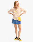 A woman stands with hands on hips, wearing a beige striped shirt, denim mini skirt, Clare Vivier's Poche handwoven leather crossbody bag with strap, and yellow sneakers with black laces, against a plain white background.