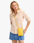 A woman stands against a plain background wearing a light pink and white striped button-up, blue denim skirt, layered necklaces, and the Clare Vivier Poche—a small handwoven leather phone bag with crossbody strap.