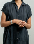 A person wearing the Raquel Allegra SS Caftan, a short-sleeve black button-up dress with subtle vertical stripes, stands with hands clasped; only the upper body is visible against a plain background.