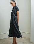 A woman stands sideways in a minimalist room, wearing the Raquel Allegra SS Caftan in black and matching sandals. Her hair is tied back, and she gazes down with a calm expression.