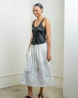 A person stands indoors by a white wall, smiling and wearing a dark sleeveless top, black sandals, and the Raquel Allegra Day Skirt—a long white skirt featuring a blue hand-painted print.