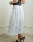 A person wears a black sleeveless top, black sandals, and a Raquel Allegra Day Skirt—a white cotton piece with subtle blue hand-painted splatters—while standing indoors on a light-colored floor.