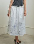 A person stands indoors on a concrete floor wearing a sleeveless black button-up top, Raquel Allegra Day Skirt with blue splatter print near the hem, black sandals, and a silver bracelet.