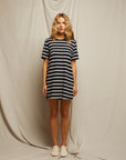A woman faces forward in front of a beige fabric backdrop, wearing the Perfectwhitetee Rhett SS Tee Dress—a relaxed, black and white striped dress—paired with light sneakers and a neutral expression.