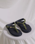 The FLO Sandals by Flowze, featuring black lightweight soles and green rope-style straps, are displayed on a round white fabric surface with a draped white cloth background.