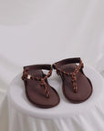 Flowze's FLO Sandals feature brown leather with knotted and braided straps, offering comfort. They are showcased on a round, white-draped table against a soft white backdrop.