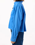 A woman with shoulder-length brown hair is shown from the side against a white background, wearing the AMO Denim Ruth Crop Shirt—an oversized, collared bright blue long-sleeve button-up—with dark blue jeans.
