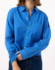 A person wears the AMO Denim Ruth Crop Shirt in bright blue with dark blue jeans, standing against a white background and holding one sleeve. Their face is partially out of frame.