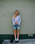 A person with long hair wearing the Ruth Oversized Shirt by AMO Denim and denim shorts stands against a green tiled wall. Their hands rest in their pockets, while a black bag sits on a distressed wooden bench in front of them.
