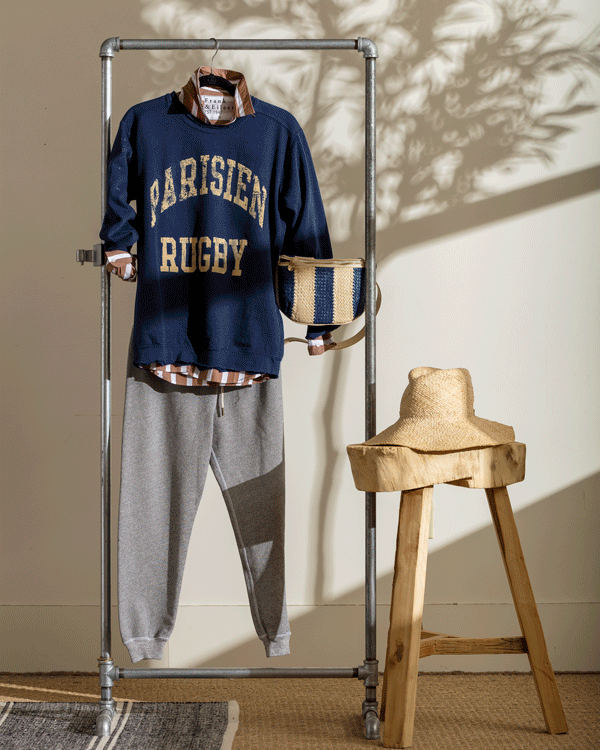 A blue rugby sweatshirt and gray sweatpants displayed on a metal rack with a tan woven handbag hanging from it, next to a rustic wooden stool with a straw hat on it, with shadow patterns on the wall.