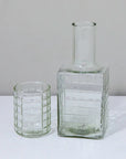 The Be Home Ruffle Glass Bedside Set—a chic clear tumbler and matching square bottle with a grid pattern—rests side by side on a white surface against a plain background.