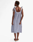 A woman with short hair and a gold necklace wears the Clare Vivier Rosalie Dress, a sleeveless linen dress with black and white gingham pattern and tied shoulder straps, paired with black sandals. She stands against a plain white background.