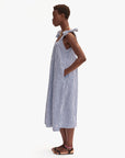 A person stands in profile wearing the Clare Vivier Rosalie Dress—a loose, sleeveless blue and white gingham linen dress with a square neckline, shoulder ties, side pockets—and black sandals. The background is plain white.