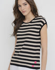 A woman with long brown hair wears the Lisa Todd Summer Lovin Sweater, a sleeveless black and beige striped top with two crochet hearts near the hem, paired with white pants, standing against a plain light background.