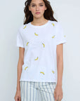 A person wears the Lisa Todd Bananas Tee, featuring small yellow banana embroidery, paired with light striped pants against a plain background for a fresh summer look.