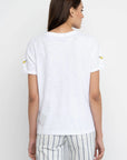 A woman with long brown hair stands with her back to the camera, wearing a Lisa Todd Bananas Tee and white pants with blue and beige stripes, creating a fresh summer look against a light background.