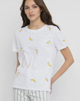 A woman with long brown hair wears the Lisa Todd Bananas Tee, a relaxed fit white T-shirt featuring small yellow banana embroidery, paired with light-colored striped pants. She stands against a plain white background, smiling slightly at the camera.