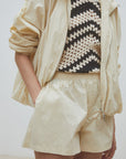 Wearing the SUNCOO Boulbi Short with an elastic waist, a light beige windbreaker, and a black-and-white crocheted zigzag top, this person channels a sporty vibe, left hand casually tucked in their shorts pocket.
