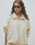 A person with long wavy hair wears black sunglasses, gold hoop earrings, and the SUNCOO Derek Jacket in light beige with matching pants, standing against a plain light background.