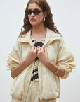 A woman in the SUNCOO Derek Jacket, matching pants, a black and white floral crochet top, black sunglasses, and gold jewelry stands against a light background. She has wavy brown hair and a neutral expression. Stylish weather protection.