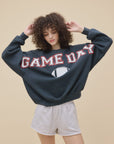 A curly-haired person wears the DayDreamer Game Day Sweatshirt, a dark fleece crew with a football graphic, paired with light gray shorts, standing against a plain beige background.