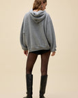 A person stands with their back to the camera in a loose DayDreamer FleetWood Mac Hoodie, black shorts, sheer black tights, and knee-high black boots against a plain beige background.