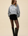 A person wears an oversized DayDreamer FleetWood Mac Hoodie, black shorts, sheer black tights, and black knee-high boots while standing against a plain beige background.
