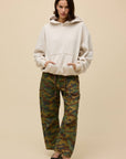 A woman stands against a beige background wearing the DayDreamer OS Hoodie, paired with camouflage pants and black ballet flats, her hands in the hoodie pocket and expression neutral.