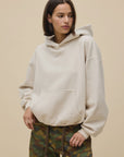 A woman models the DayDreamer OS Hoodie, an oversized beige style, paired with camouflage pants. She stands against a plain beige background, gazing slightly left with one hand in her pocket.