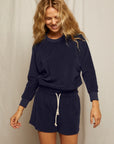 A woman with wavy blonde hair smiles while wearing the Perfectwhitetee Saylor Loop Terry Sweatshirt in navy blue and matching shorts with a white drawstring, standing before a light background.
