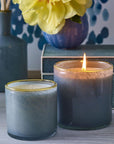 Two blue Beach House candles by Lafco NY in hand-blown glass sit side by side on a table; one glows while the other is unlit. A yellow flower in a vase and a decorative book or box evoke the Sea & Dune-inspired ambiance.