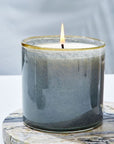 A lit Beach House Candle by Lafco NY sits in a glossy gray hand-blown glass vessel with a gold rim on a round marbled surface, set against a soft white background.