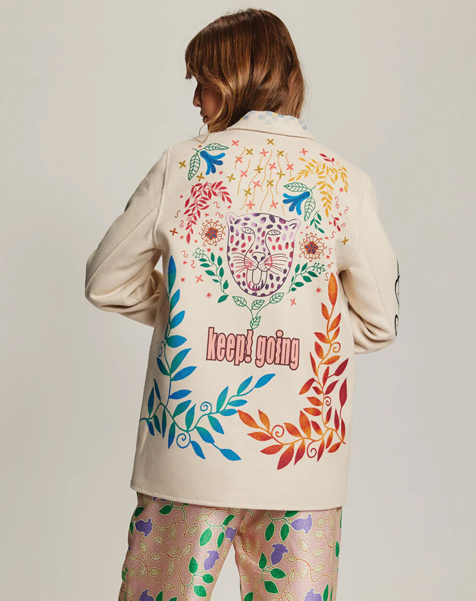 A person with shoulder-length hair faces away, wearing the Me369 Juliana Wool Jacket—off white with colorful florals, a pink tiger face, and "keep! going" on the back—paired with shimmery multicolored pants.