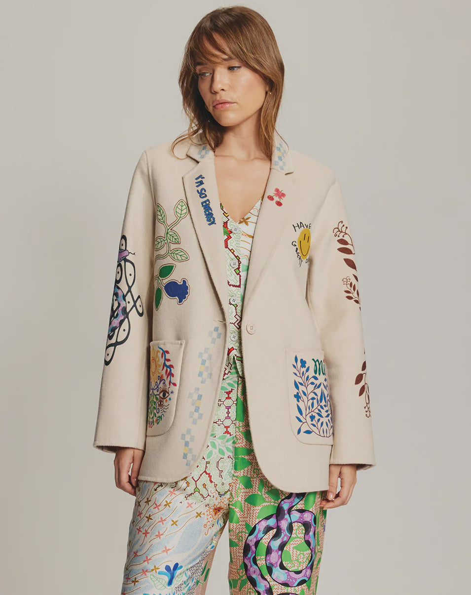 A woman wears the Me369 Juliana Wool Jacket, an off-white piece featuring vibrant, whimsical embroidery, paired with matching patterned pants and top, standing against a plain background.