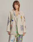 A woman wears the Me369 Juliana Wool Jacket, an off-white piece featuring vibrant, whimsical embroidery, paired with matching patterned pants and top, standing against a plain background.