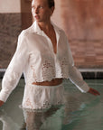 A woman stands waist-deep in water, wearing Cissa's The Shorty Shirt—a white, long-sleeved men’s shirt with matching embroidered shorts. She gazes to the side in a serene indoor space with stone and tile walls.