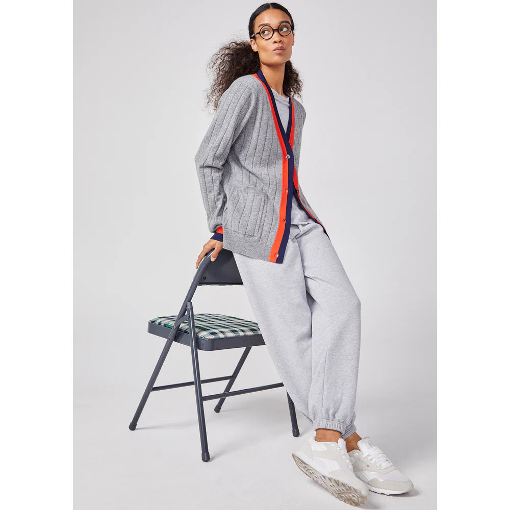 A person with curly hair and glasses sits on a folding chair, wrapped in a cozy gray cardigan with red trim over matching joggers. They sport white sneakers against a plain white background, effortlessly styled in Kule's The Women's Organic Sweatpants for ultimate comfort.