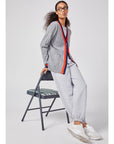 A person with curly hair and glasses sits on a folding chair, wrapped in a cozy gray cardigan with red trim over matching joggers. They sport white sneakers against a plain white background, effortlessly styled in Kule's The Women's Organic Sweatpants for ultimate comfort.