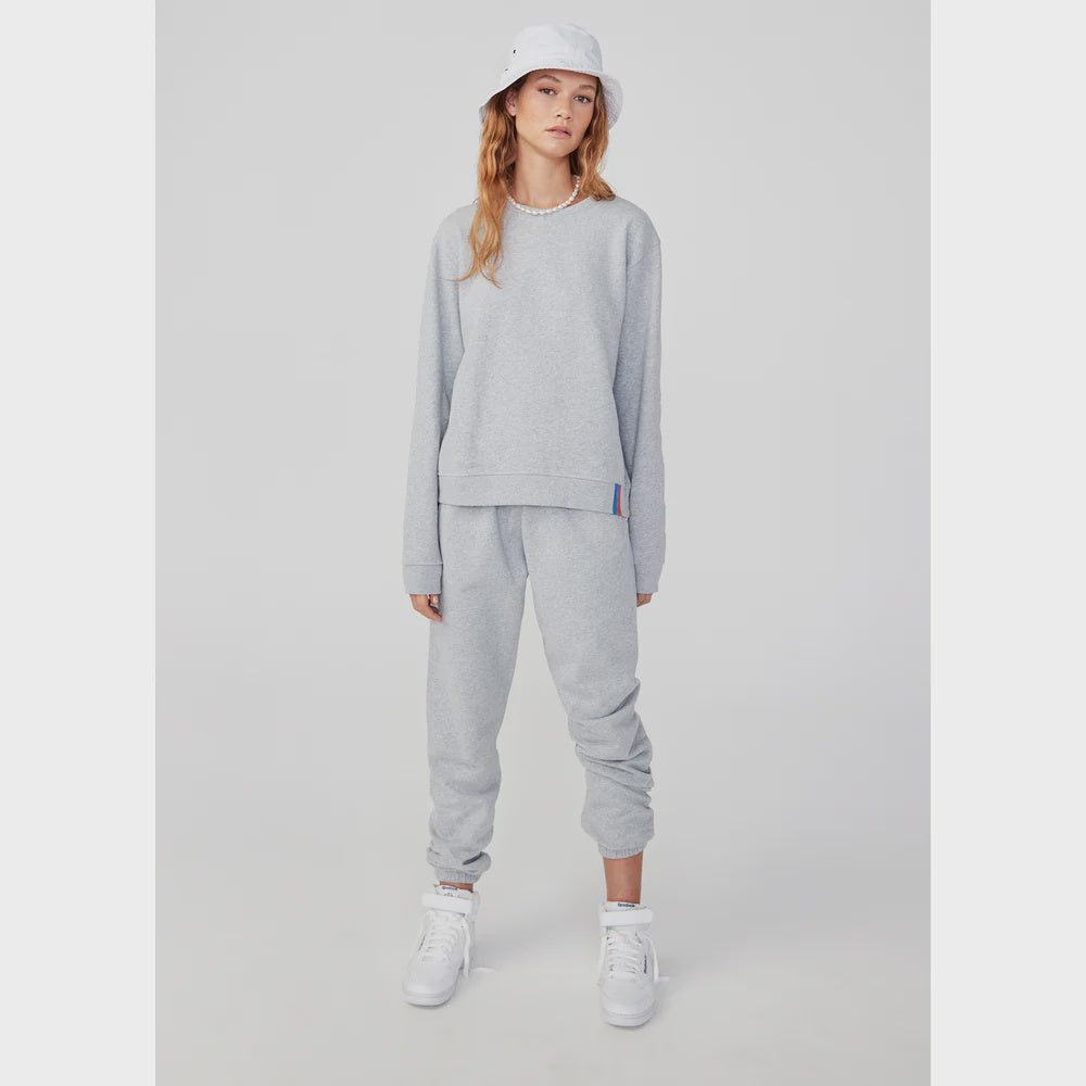 A person stands against a plain background in Kule's Women's Organic Sweatpants and a light gray sweatshirt. Complementing their relaxed pose are white sneakers and a white bucket hat, enhancing the laid-back vibe. The outfit is practical and stylish, being entirely machine washable.