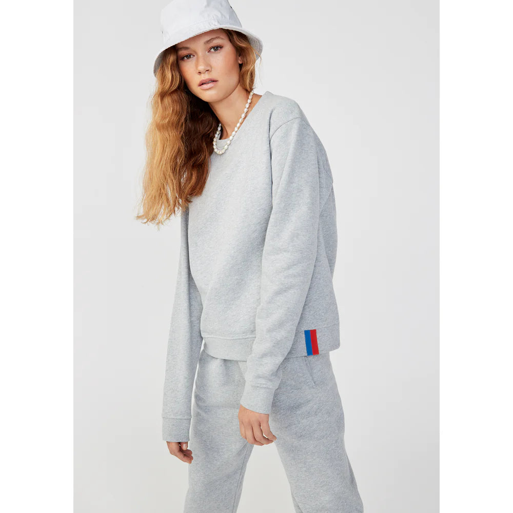 A person with long hair wears a white bucket hat, a light gray leisure tuxedo, and The Women's Organic Sweatpants from Kule. These pants feature a small red and blue tag on the side, adding elegance as they pose against a plain background.