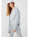 A person with long hair wears a white bucket hat, a light gray leisure tuxedo, and The Women's Organic Sweatpants from Kule. These pants feature a small red and blue tag on the side, adding elegance as they pose against a plain background.