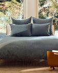 A sunlit bedroom features a neatly made bed with the Coyuchi Inc Toro Canyon Org. Duvet Cover in Marine King, four pillows, wooden side tables, a lamp, and a magazine holder by large windows overlooking trees.