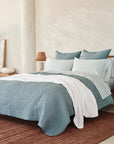 A modern bedroom with a bed covered in blue and white 100% organic cotton bedding, Coyuchi Org Percale Pillowcase Dew Chrysanthemum S/2 Queen, two wooden nightstands with woven lamps, brown rug, potted plant, and neutral wall decor in a bright space.