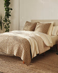 A cozy bedroom with a neatly made bed featuring the Blossom Hand Stitched Quilt by Coyuchi Inc, pillows, a cream headboard, plants, wooden furniture, a large window with greenery outside, and a woven rug on the floor.